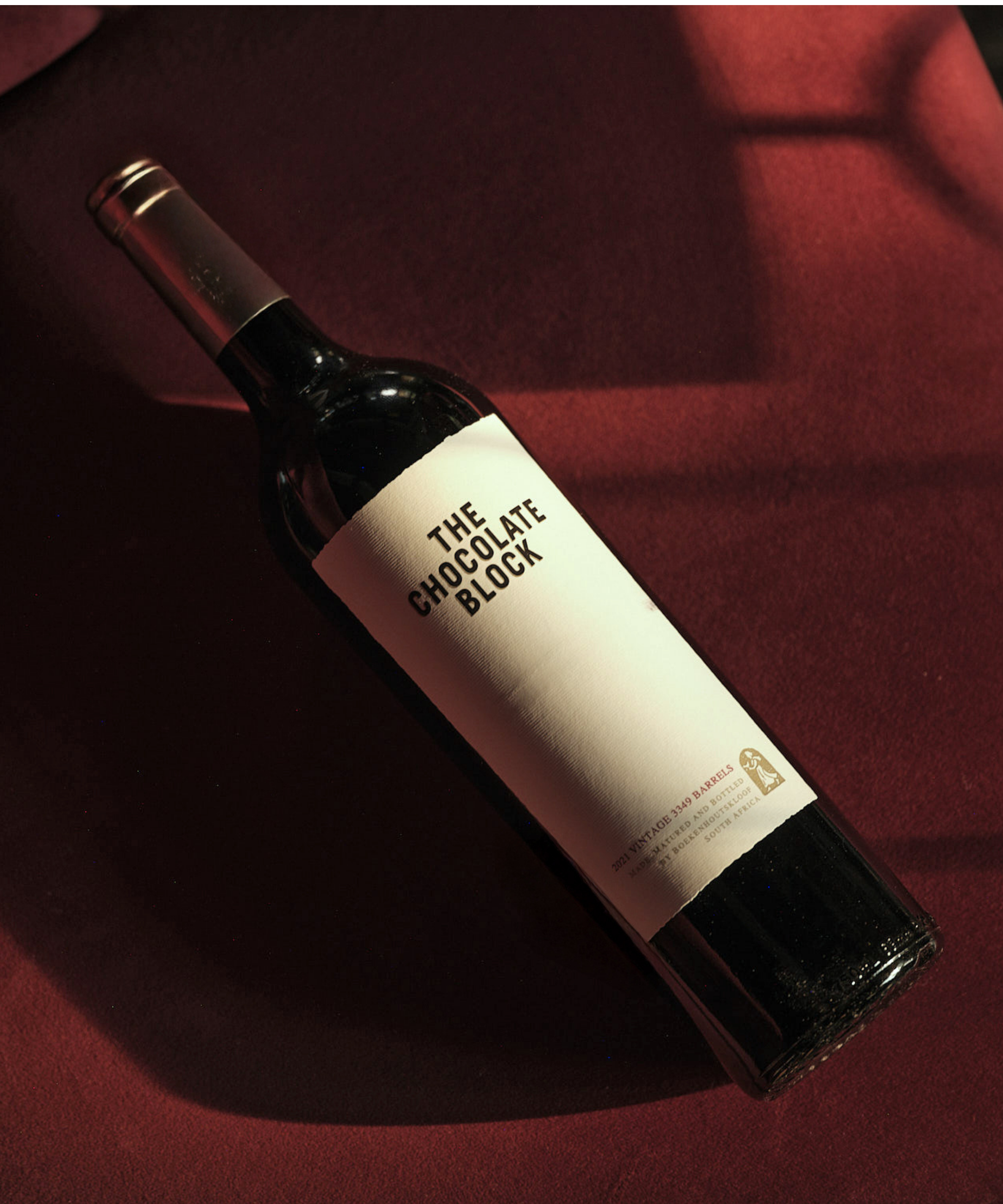 A clear glass bottle of The Chocolate Block wine resting on a dark red textured surface. The bottle's label is partially visible, showing black and white text and a small logo at the bottom.