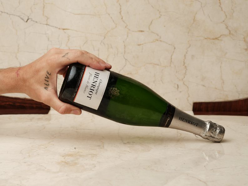 A hand with tattoos holding a tilted bottle of champagne on a marble table, with a cracked wall background.