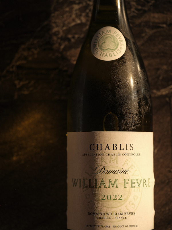 A bottle of William Fevre Domaine Chablis 2022 wine against a dark wooden background.