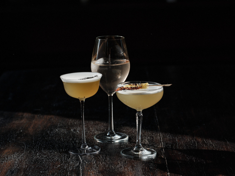 Three cocktails on a dark wooden surface, two in coupe glasses and one in a wine glass, with dark background.