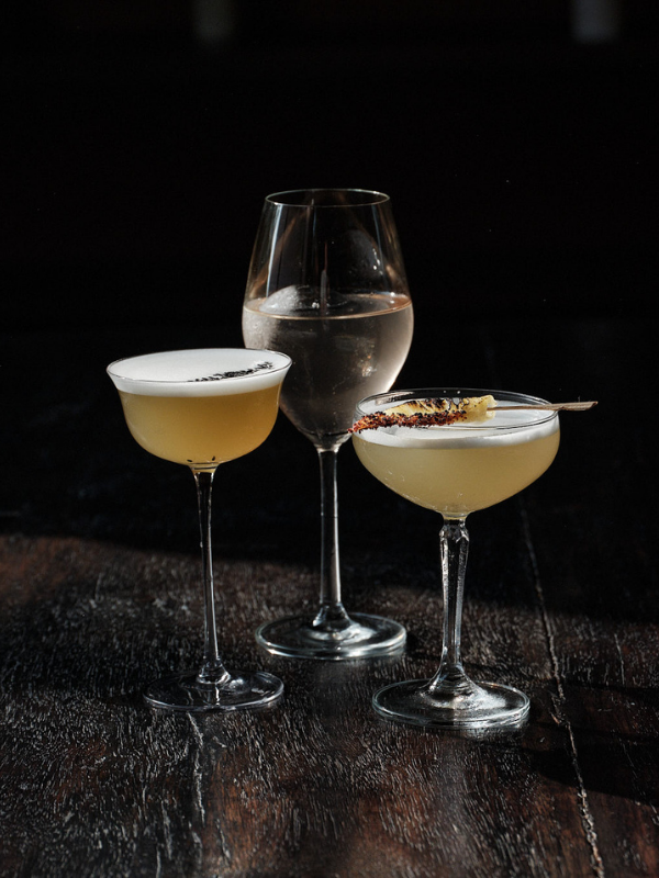 Three cocktail glasses on a dark wooden surface, with two containing light-colored drinks and one filled with water or clear liquid, one cocktail garnished with a skewer and a salted rim.