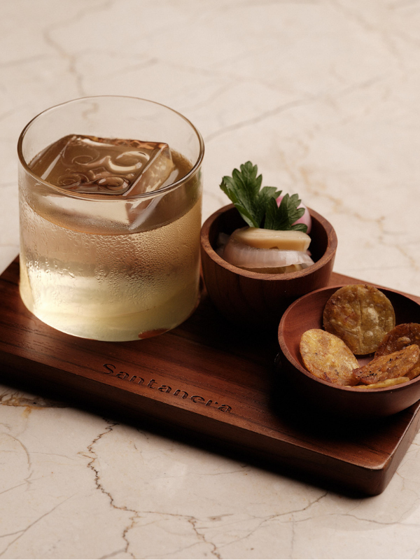 A whiskey cocktail with a large ice cube, served on a wooden tray with a small bowl of pickled onions and herbs, and a small bowl of potato chips, all on a marble countertop.