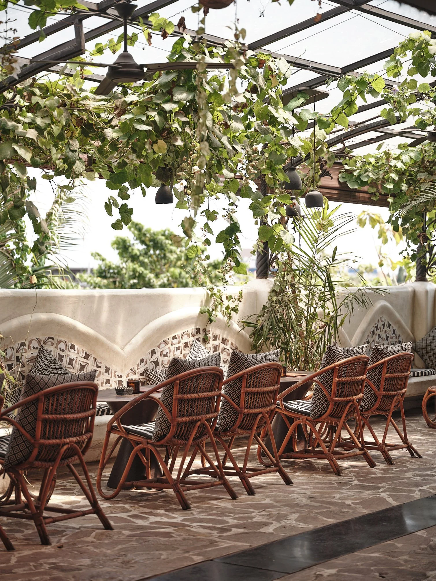 Outdoor seating area with multiple wooden chairs with patterned cushions, a stone-tiled floor, and a decorative white wall with an arched design. The space is shaded by a metal and glass ceiling covered with climbing green vines and hanging black pendant lights.