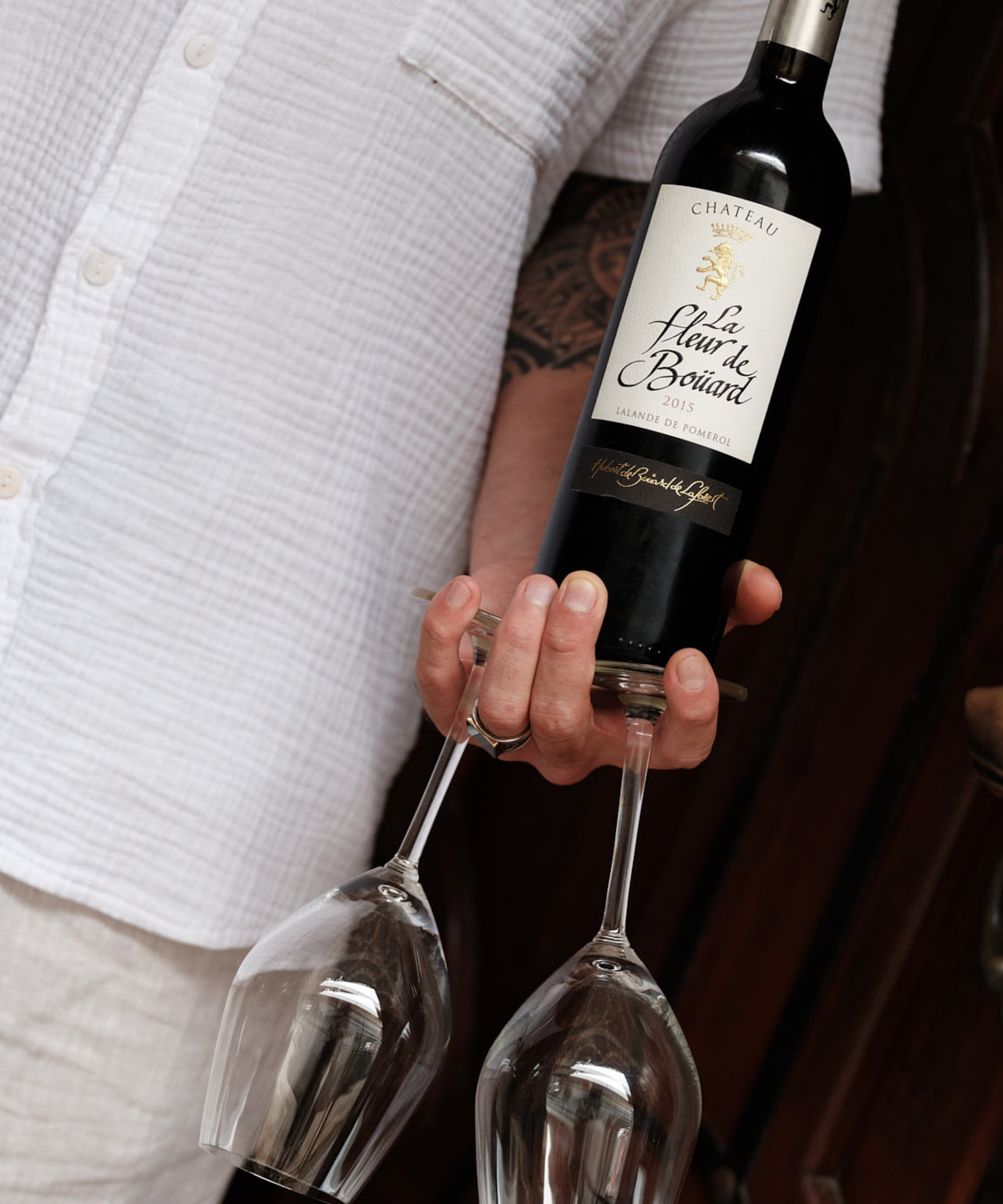 Staff member holding a bottle of red wine and two wine glasses beside the entrance to rooftop bar in Canggu, Bali.