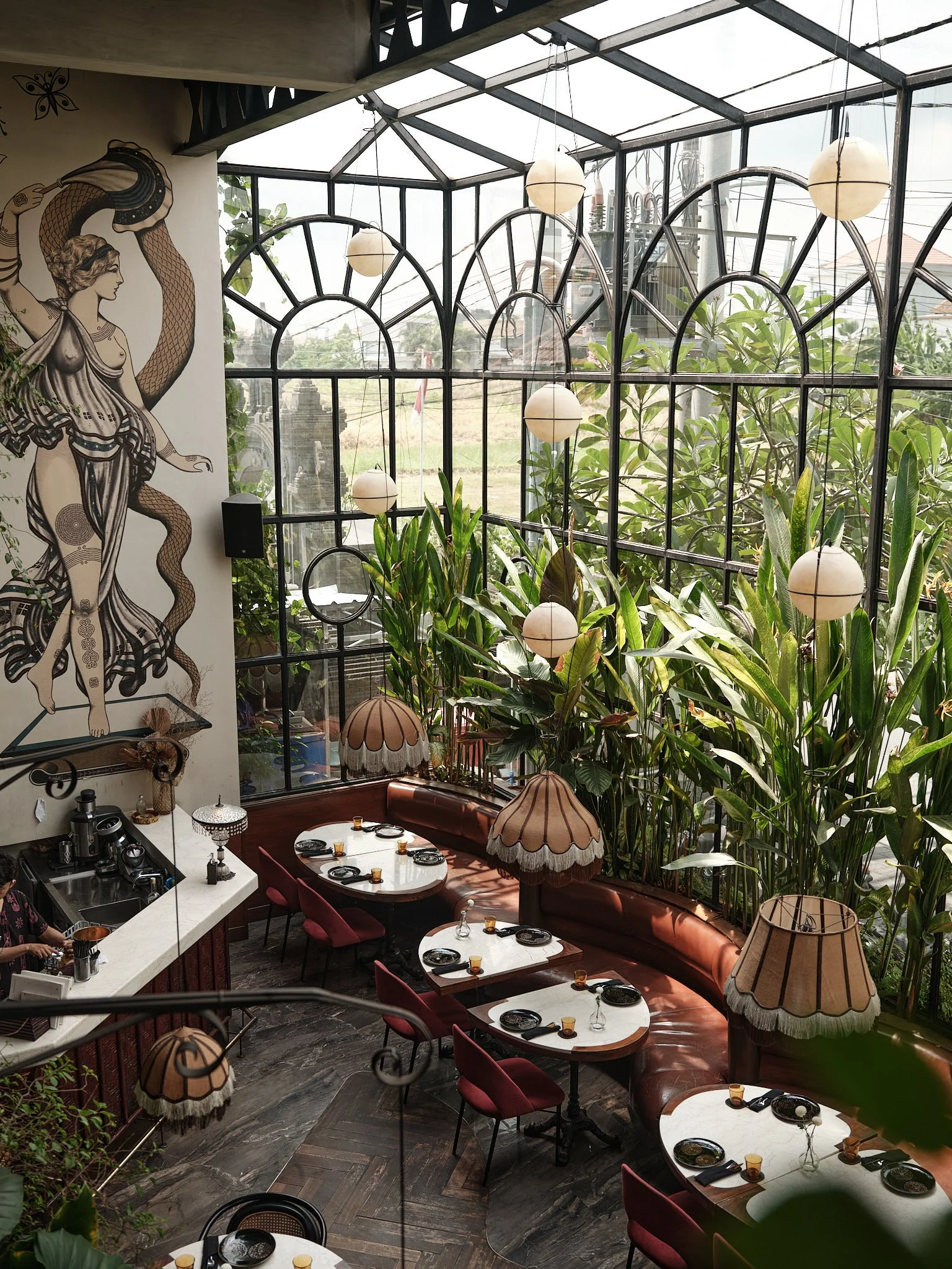 An indoor restaurant with a curved glass wall filled with green plants. There are round tables set with black plates and yellow glasses, surrounded by red chairs. Hanging lamps with beige shades adorn the space, and a mural of a woman with a snake is painted on the wall.