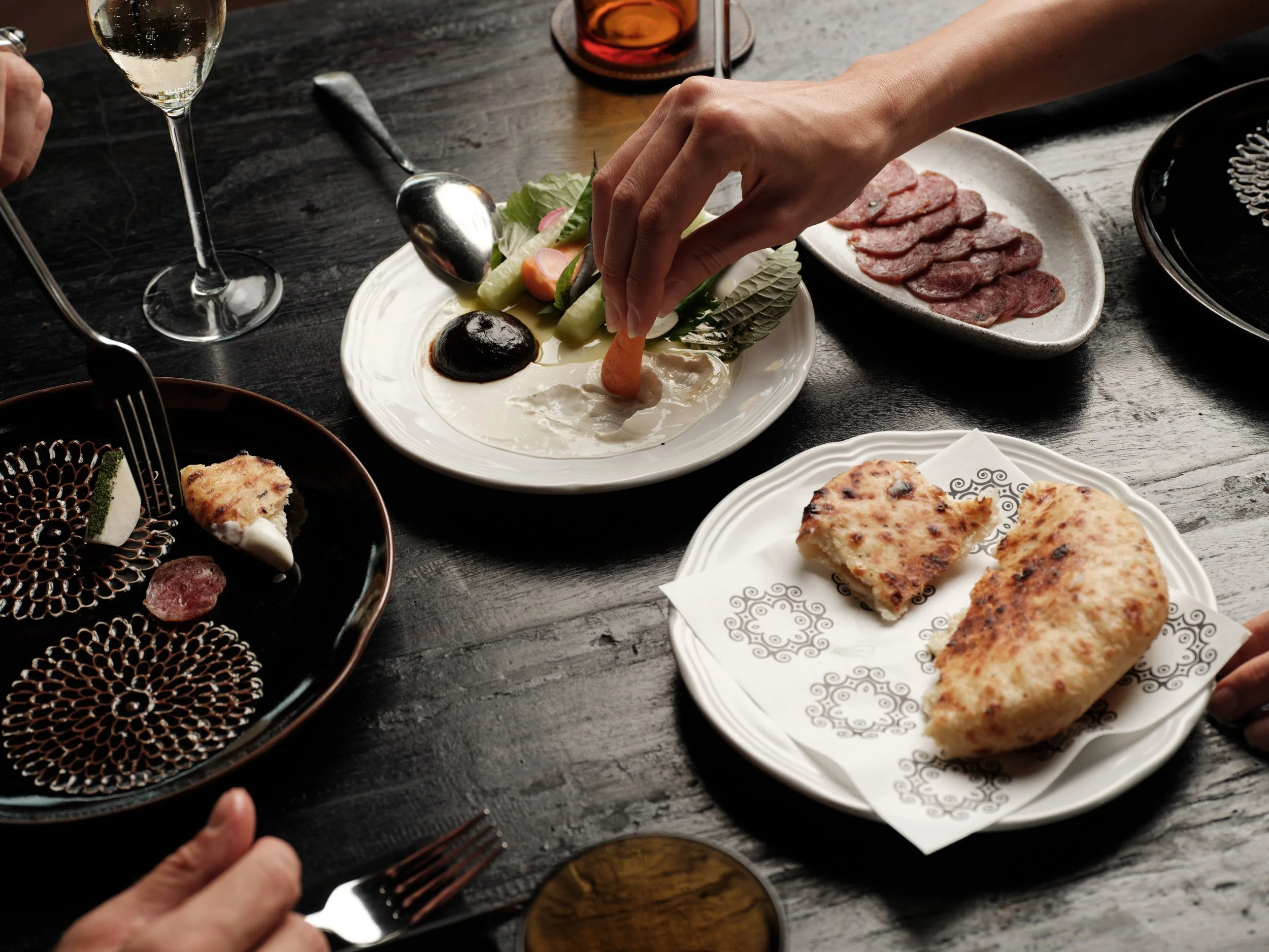 A person is serving salad with cucumbers, radishes, lettuce, and dressing on a white plate. There are slices of cured meat on a gray dish, bread on a white paper-lined plate, a glass of white wine, and a glass of pink-colored drink on the table, whic