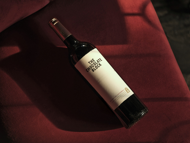 A bottle of red wine labeled "The Chocolate Block" lying on a dark red surface with shadows in the background.