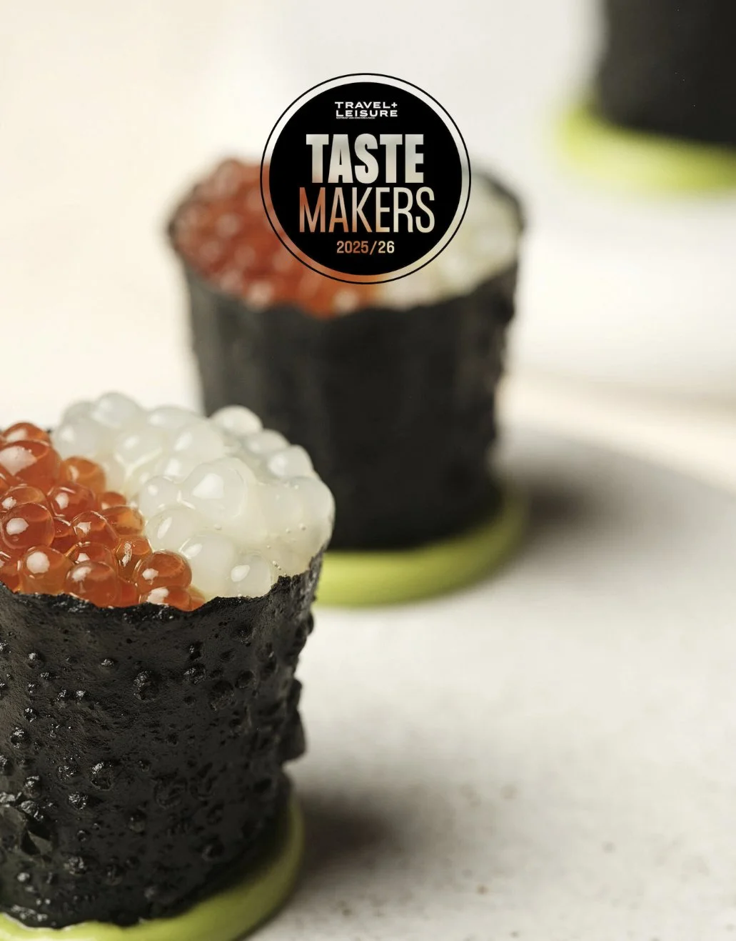 Close-up of two sushi rolls topped with white and orange fish roe, with a blurred background and a black circular logo reading 'Travel + Leisure Taste Makers 2025/26' placed on the front sushi roll.