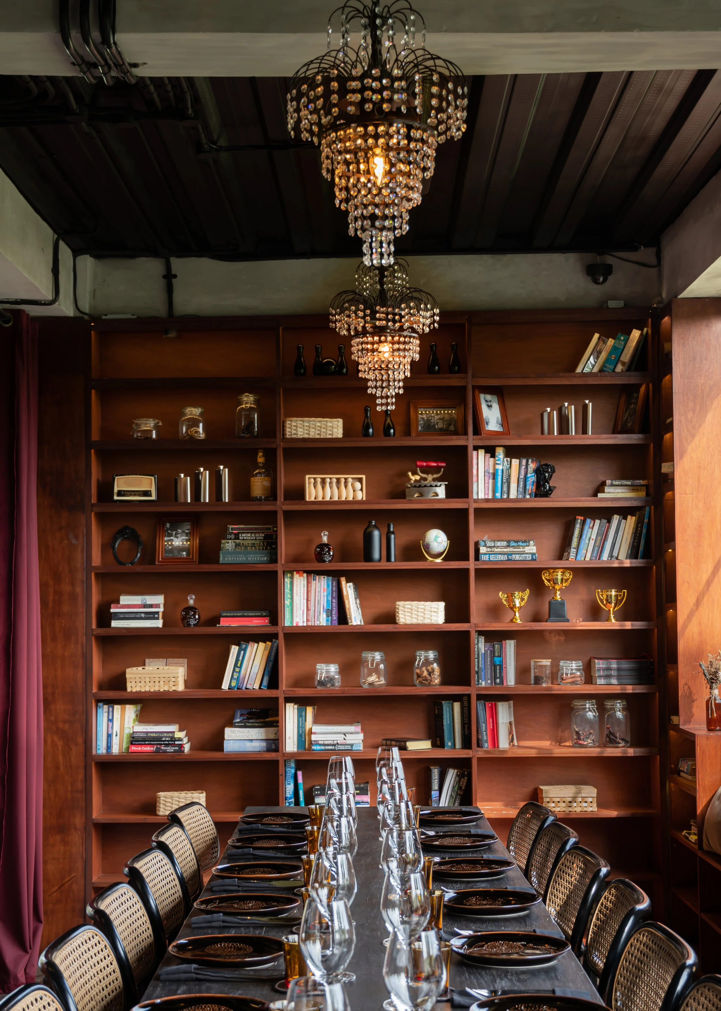 A long dining table set for a meal in front of a large wooden bookshelf filled with books, decorative items, and trophies. A chandelier with hanging crystals illuminates the room from the ceiling.