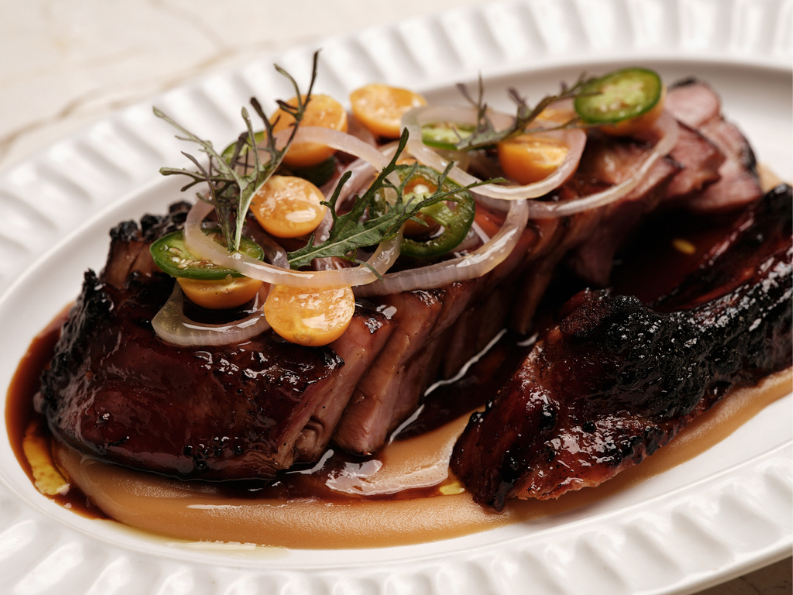 Barbecue beef brisket with onion slices, jalapeño slices, corn, and herbs, served on a white plate with sauce.