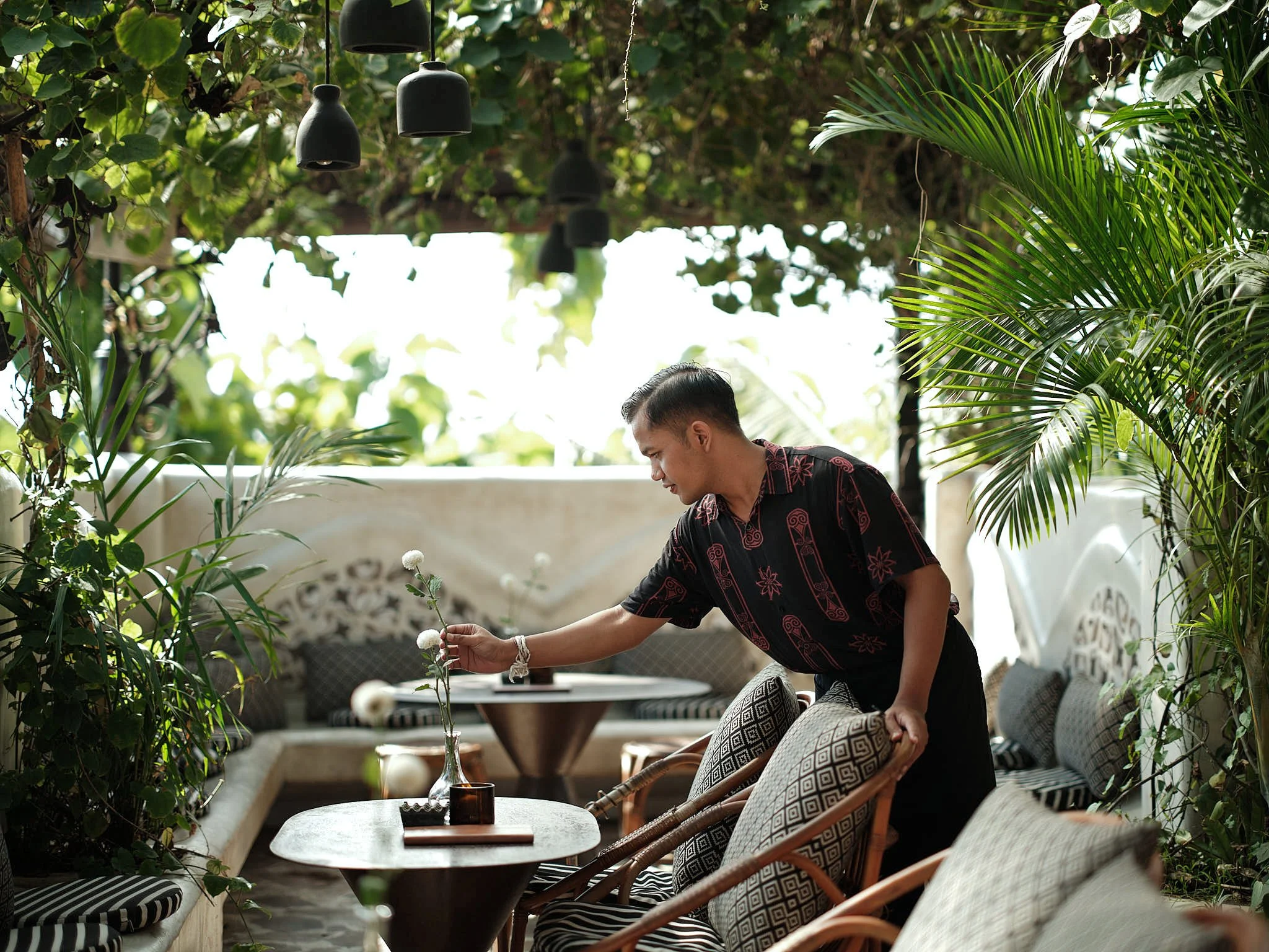 A man with dark hair and a patterned black and red shirt is arranging a flower in a tall glass vase on a table in an outdoor seating area surrounded by lush green tropical plants and trees.