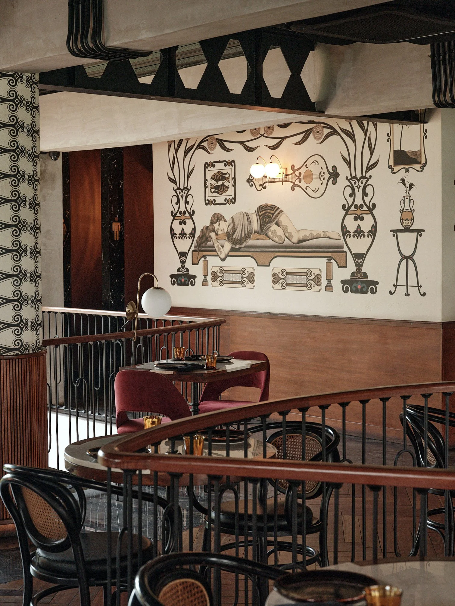 Interior of a restaurant or café with black and beige chairs, round tables set with glasses, a large art mural of a reclining woman, and decorative wall elements.