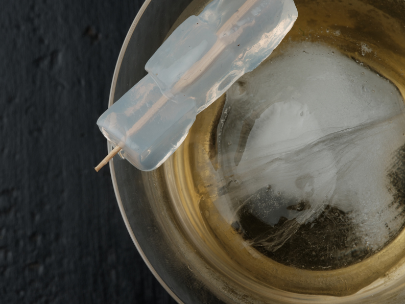 An ice cube on a skewer resting on a reflective metal surface.