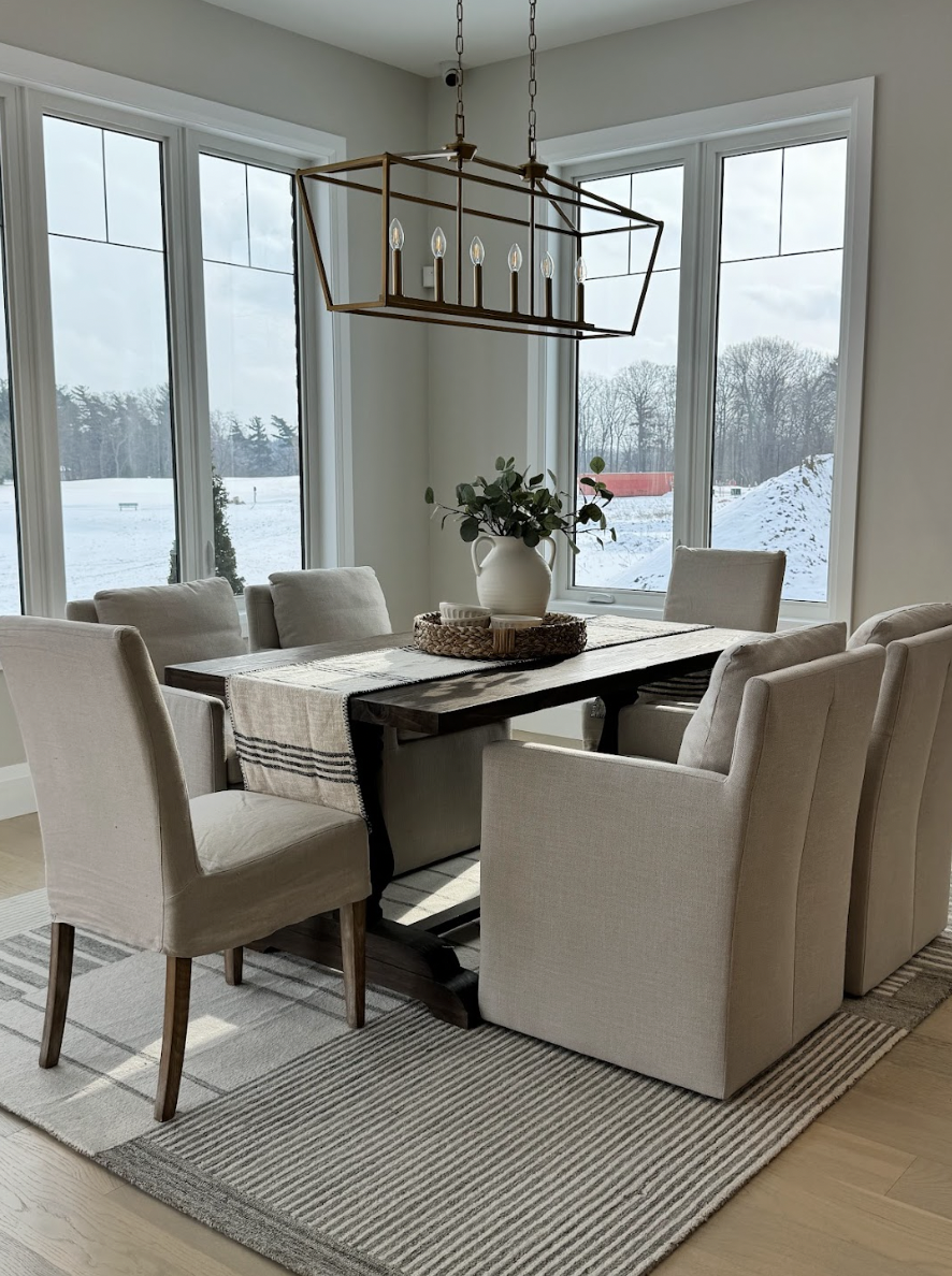 Dining room with a wooden table surrounded by six beige upholstered chairs, a striped table runner, a white ceramic pitcher with greenery, a woven tray, and a chandelier with exposed bulbs hanging above. Large windows show a snowy outdoor landscape.