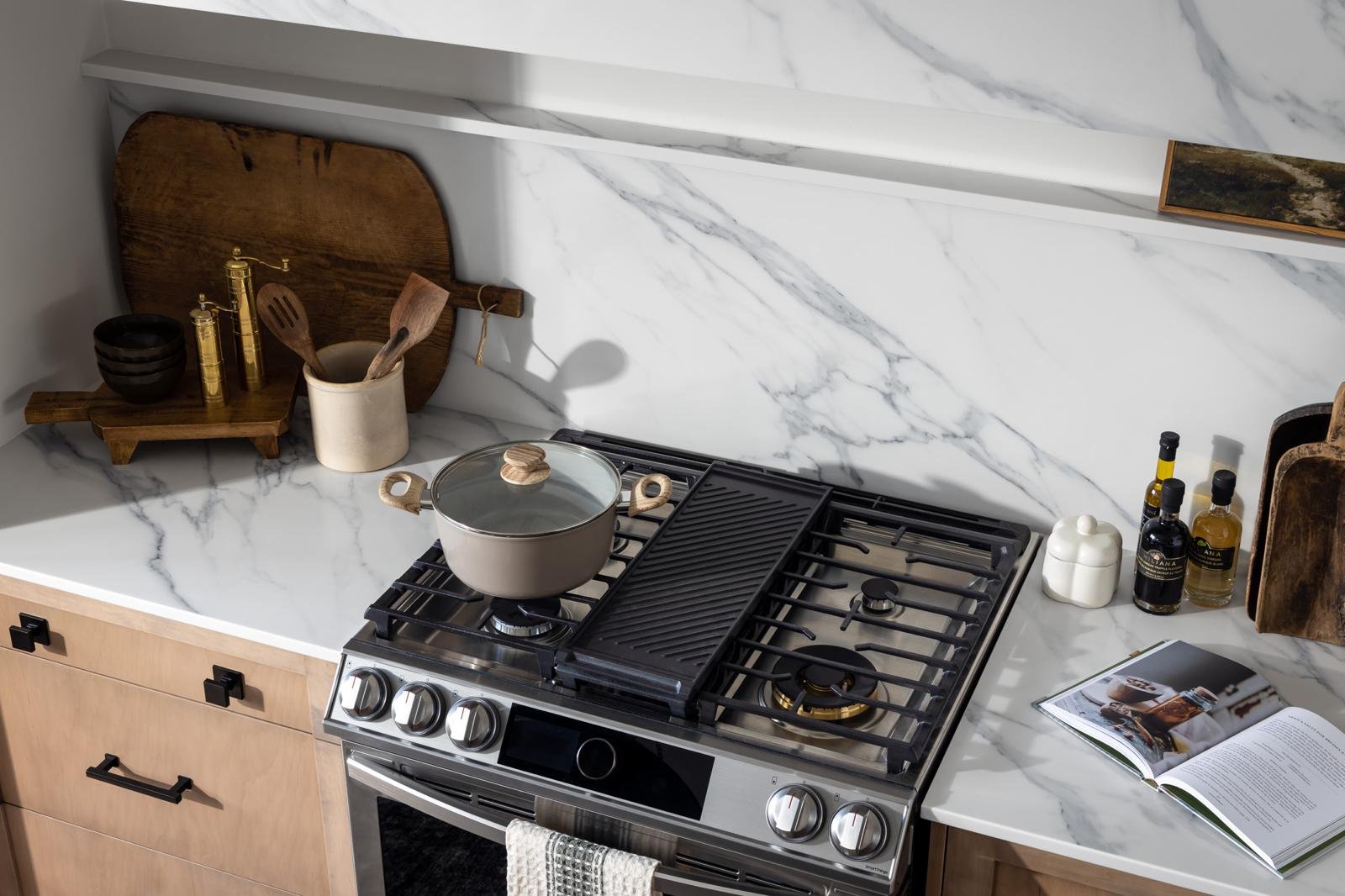A modern kitchen with a marble countertop and backsplash, wooden cabinets, and a stainless steel gas stove. The countertop has a pot with a lid on the stove, a cookbook, and bottles of oil and seasoning. There are kitchen utensils in a container, wooden cutting boards, and black bowls on the counter.