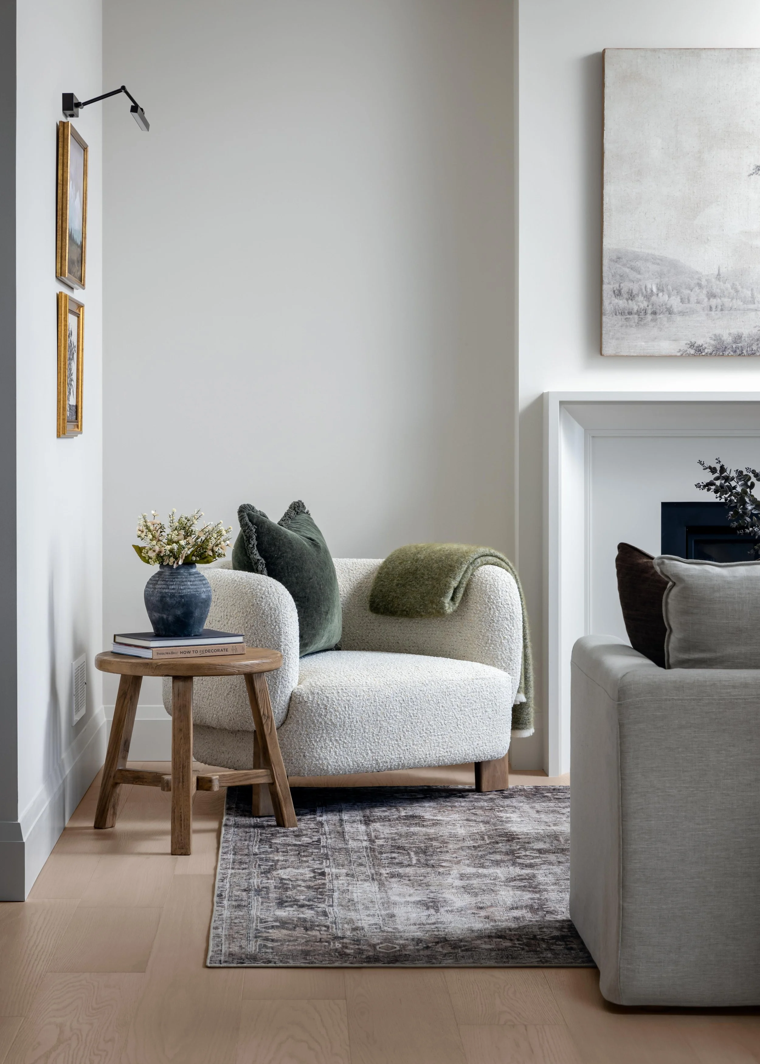 A cozy corner of a living room with a white textured armchair, green pillows, a wooden side table with a blue vase and books, a patterned rug, and a white wall with framed pictures and a wall-mounted lamp.