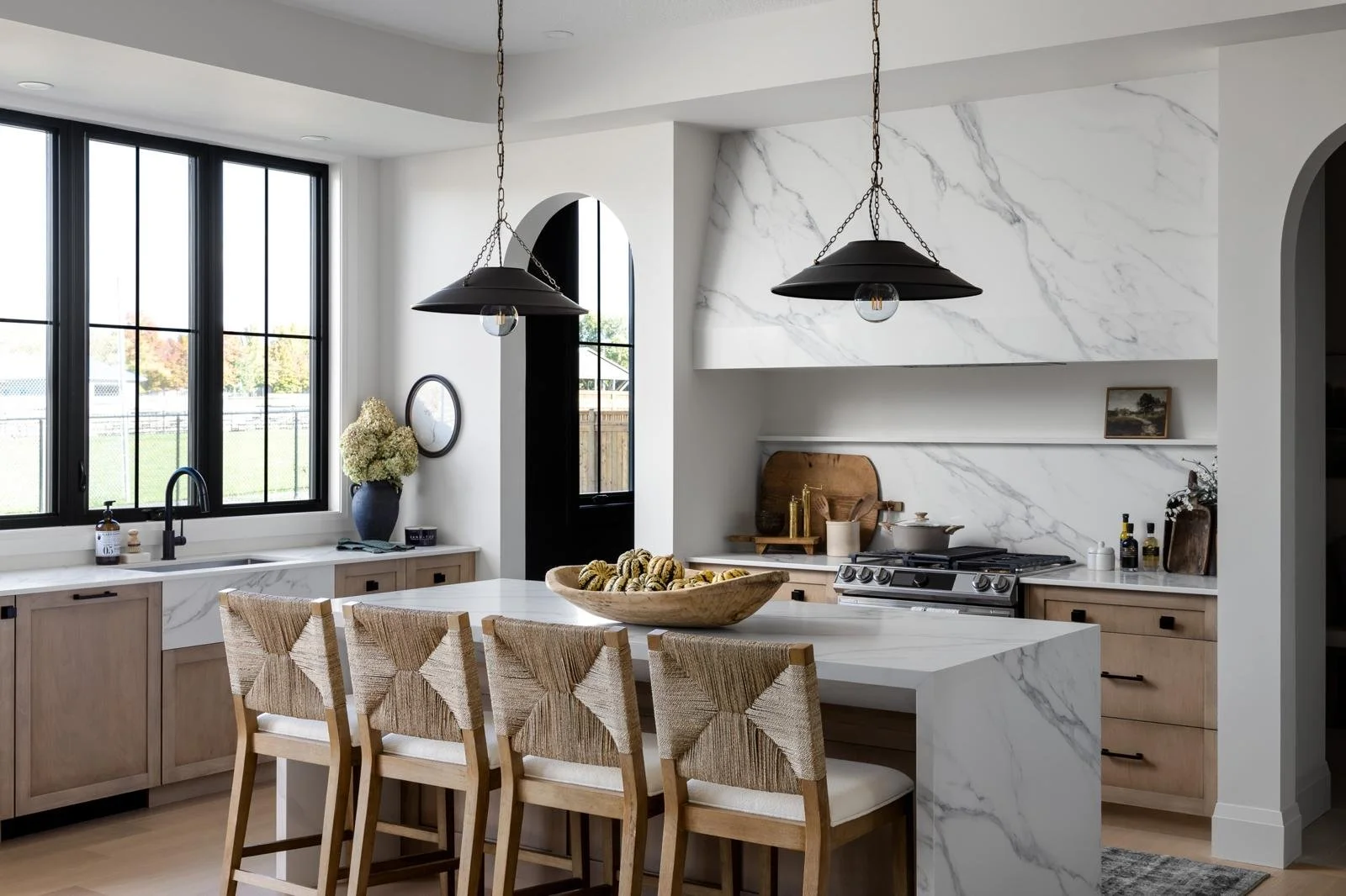 Modern kitchen with white marble countertops and backsplash, light wood cabinets, black fixtures, large window, and woven bar stools at an island with a wooden bowl of snacks.