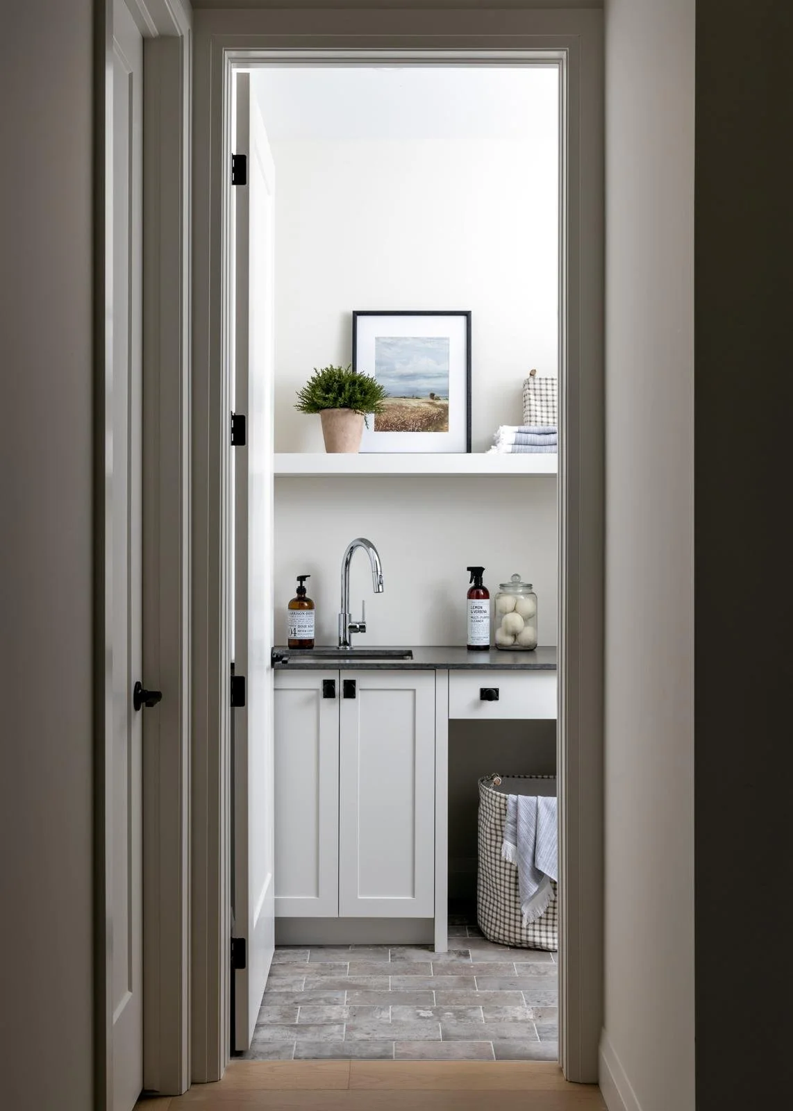 A small laundry room with white cabinets, a gray countertop, a silver faucet, framed artwork, a potted plant, folded towels, soap dispensers, a jar of cotton balls, and a laundry basket with a towel.
