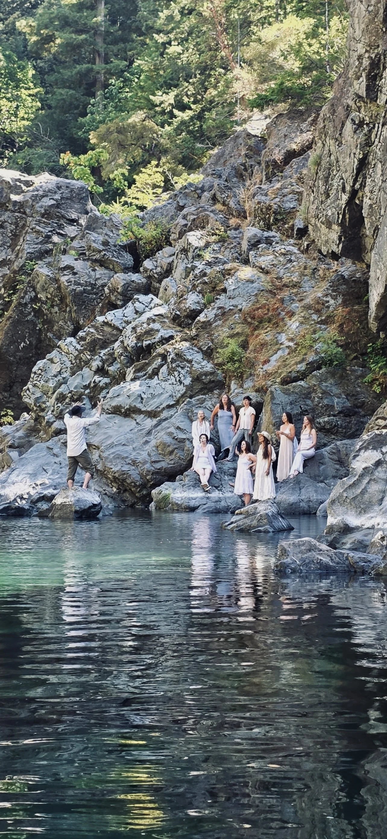A group of women dressed in white and light-colored clothing posing on rocks beside a river in a forested area, with a man taking their photo.