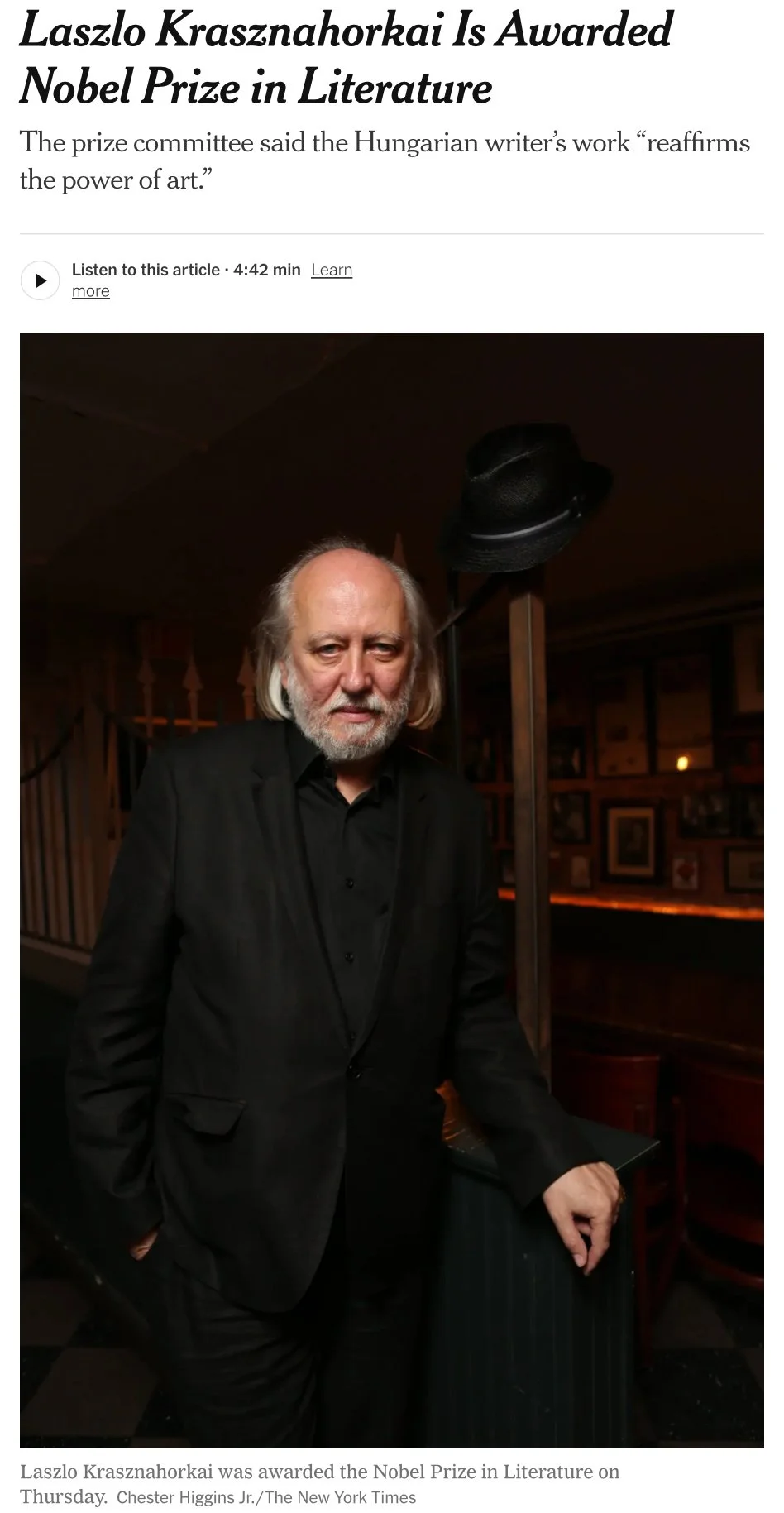 A man with long gray hair and a beard wearing a black suit, standing indoors. There is a hat hanging on a stand behind him, and framed pictures on the wall in the dimly lit background.
