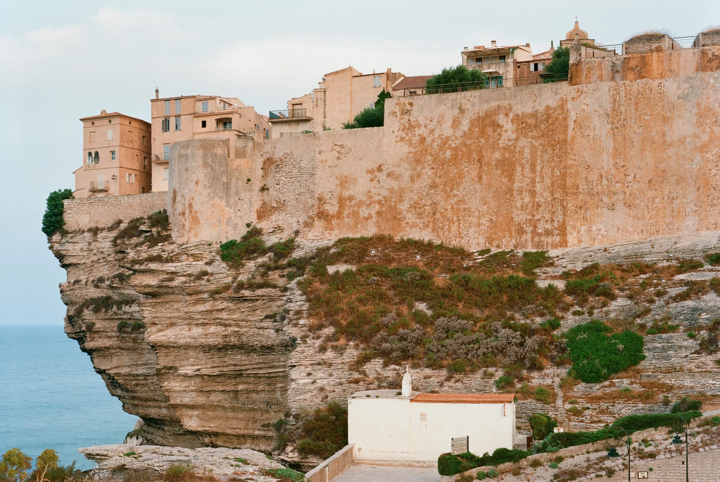 Cliffside buildings and walls overlooking the sea, with some greenery and a small white structure at the bottom.
