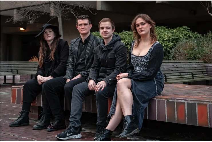 Four young adults sitting on a brick ledge outdoors with trees and a bench behind them, dressed in dark clothing.