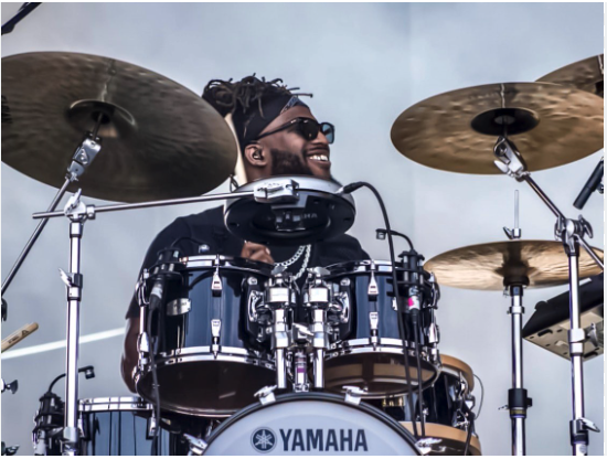 A smiling man with dreadlocks and sunglasses playing a Yamaha drum set.