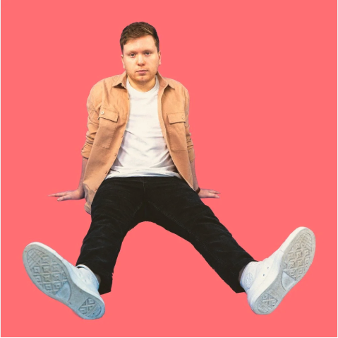 Young man sitting on the floor with legs spread apart, wearing a beige jacket, white t-shirt, black pants, and white sneakers, against a pink background.