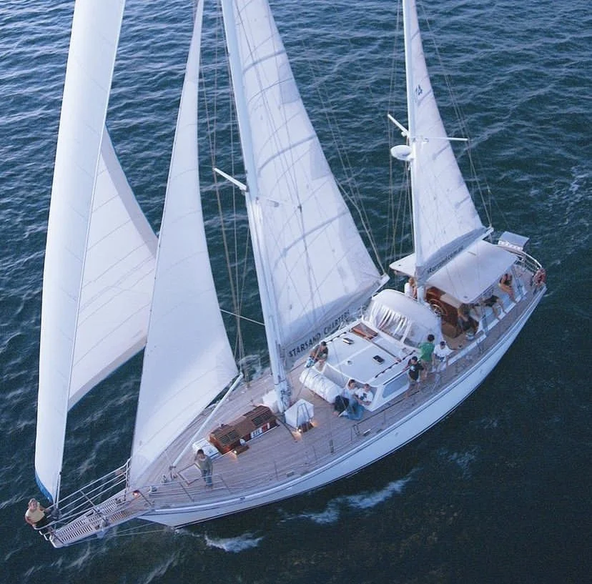 An aerial view of a white sailing yacht with multiple sails on the water, with several people on deck.