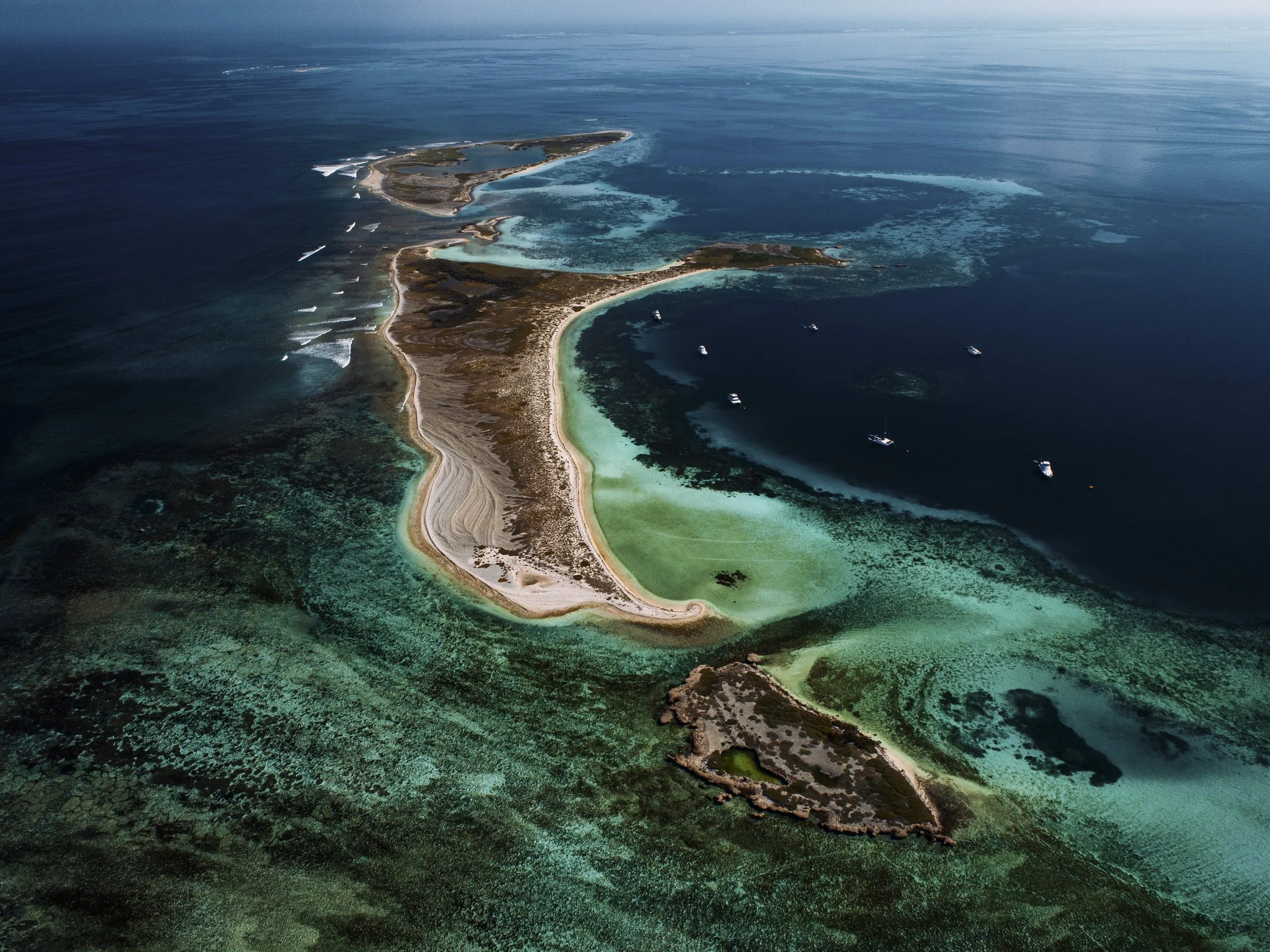 An aerial view of a coastal island with sandy beaches, clear turquoise waters, and boats anchored around it.