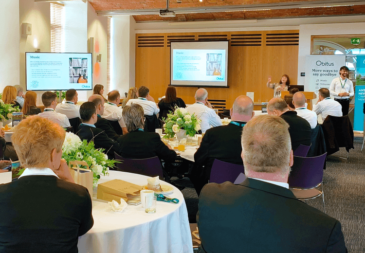 People attending a conference or seminar in a large room with round tables, presentation screens, and floral centerpieces.