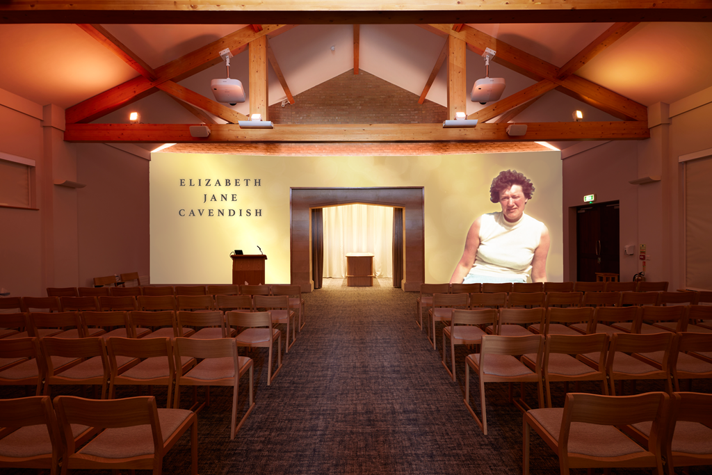 A conference room with wooden beams on the ceiling, rows of empty chairs facing a stage with a projector screen displaying the names Elizabeth, Jane, and Cavendish, and a large photo of a woman on the screen.