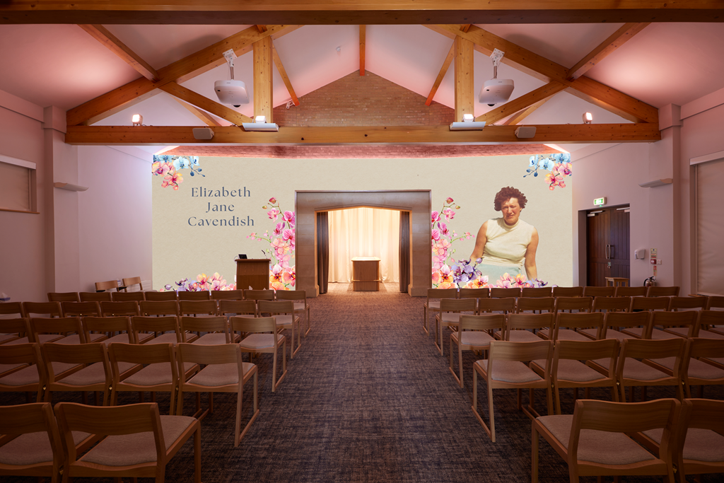 Indoor event space with wooden ceiling beams, rows of chairs facing a stage with a large screen displaying the name Elizabeth Jane Cavendish and a woman in a sleeveless dress, surrounded by floral graphics.