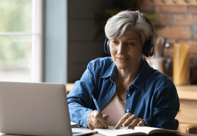 Elderly woman with gray hair wearing headphones, sitting at a table with a laptop, writing in a notebook in a cozy, well-lit room.
