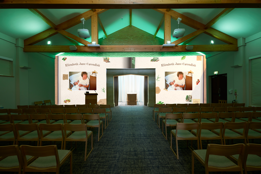 A theater with a stage featuring a large screen displaying photos of a woman holding a baby and the name Elizabeth Jane Cavendish, with rows of wooden chairs facing the stage.