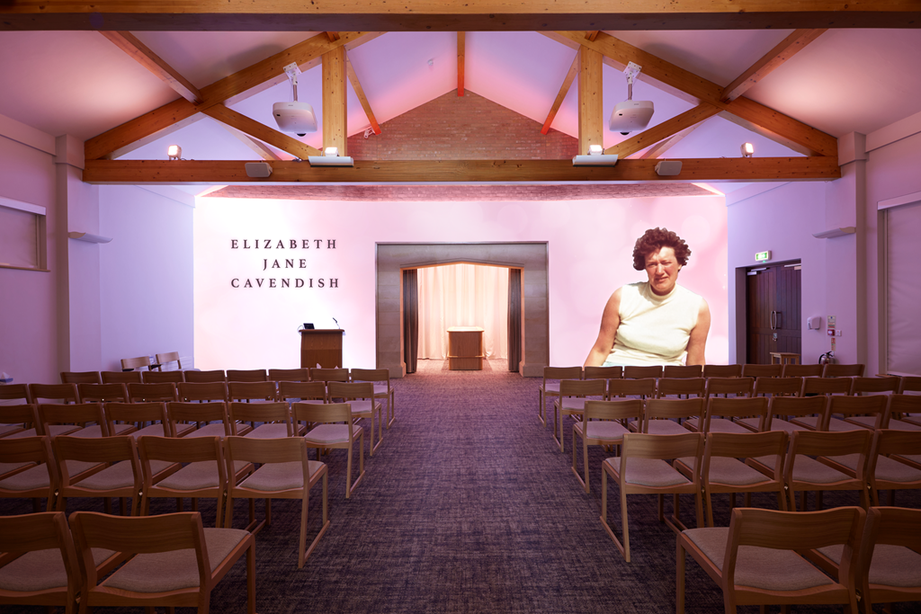 A small auditorium with rows of wooden chairs, a podium at the front, and a large projected image of a woman on the right side. The wall behind the stage has the text 'Elizabeth Jane Cavendish'.