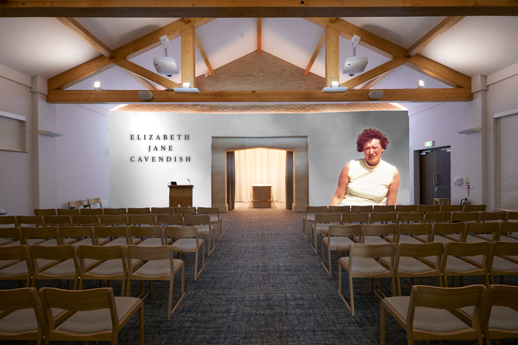 Empty auditorium with wooden chairs, a stage with a podium, and a large screen displaying an older woman with curly red hair. The wall behind the stage has the name 'Elizabeth Jane Cavendish' in black letters.