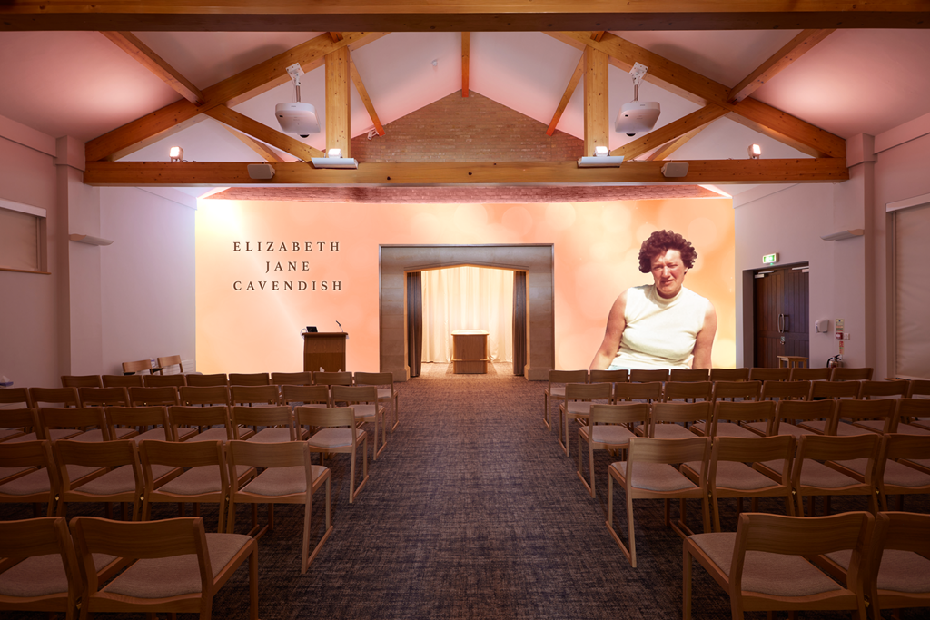 An empty event hall with wooden chairs arranged in rows, a presentation area with a lectern, a large screen displaying the name Elizabeth Jane Cavendish and a woman speaking, and exposed wooden beams on the ceiling.