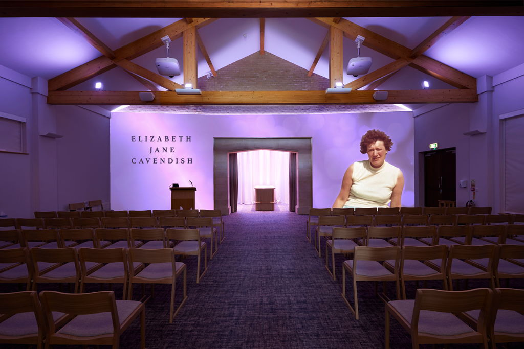 Empty conference room with rows of wooden chairs, a small podium, and a stage with a curtain. The back wall displays the name Elizabeth Jane Cavendish. A large projection of a woman with short curly hair and a sleeveless white top is on the right side of the room.