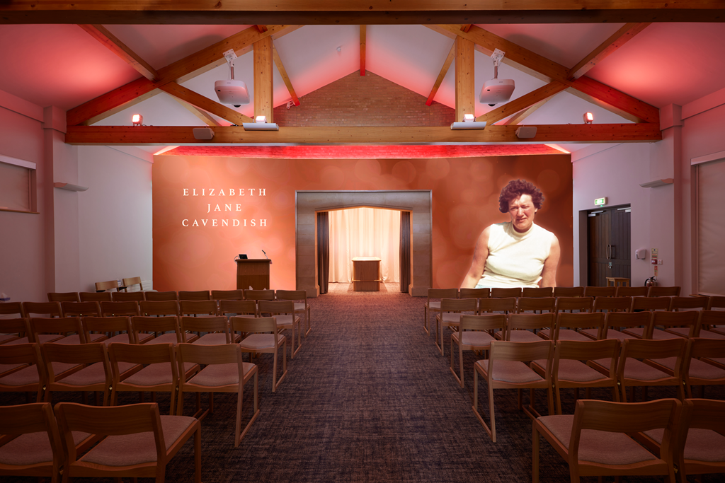 A presentation hall with rows of empty wooden chairs, a stage at the front with a podium, and a large screen displaying a woman’s face. The backdrop of the stage features the name Elizabeth Jane Cavendish.