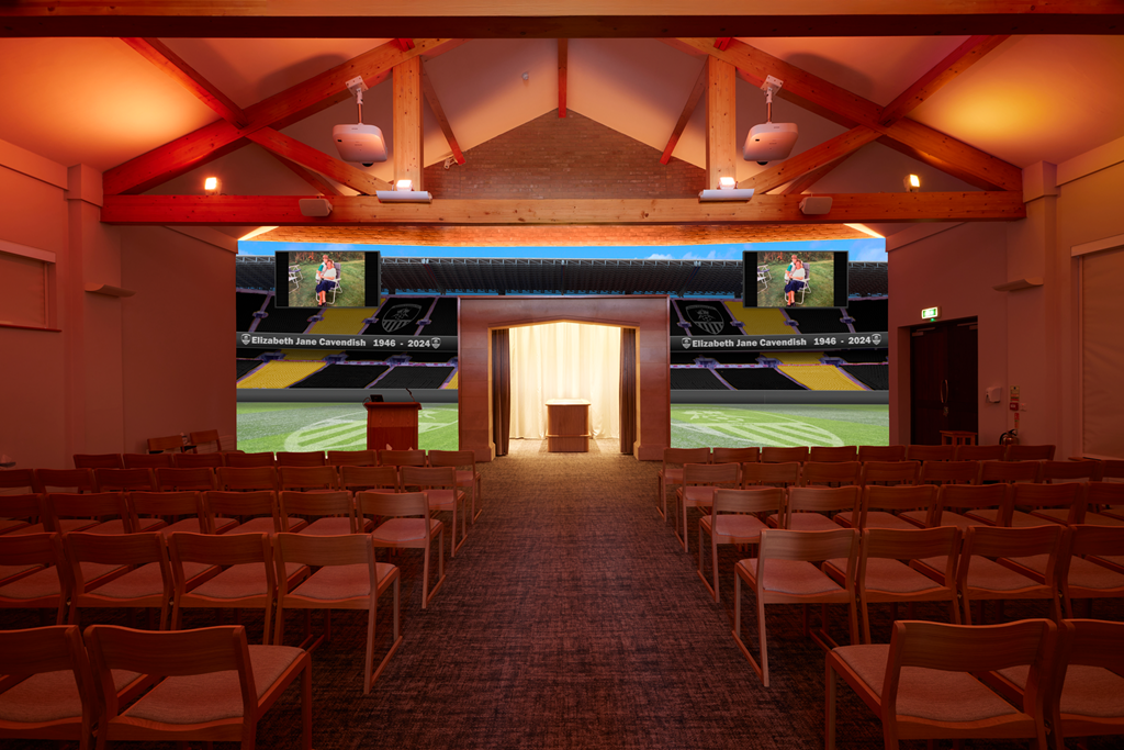 Indoor memorial service room with rows of chairs facing a stage and two screens displaying an image of a woman in a park, with a scoreboard in the background honoring Elizabeth Jane Cavendish 1946-2024.