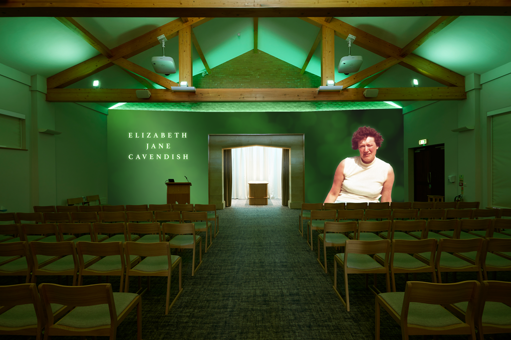 An auditorium with wooden chairs facing a stage, a green backdrop with white text reading 'Elizabeth Jane Cavendish,' and a projected image of a woman with curly hair in a white sleeveless top on the right side of the stage. There is a small table and a podium on the stage, and the space has wooden beams on the ceiling with green lighting.
