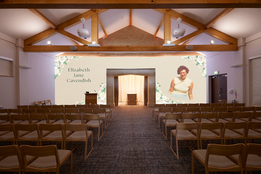 An empty wedding ceremony space with chairs facing a large screen displaying the name Elizabeth Jane Cavendish and a photograph of a woman with curly hair. The room has wooden beams and a high ceiling.