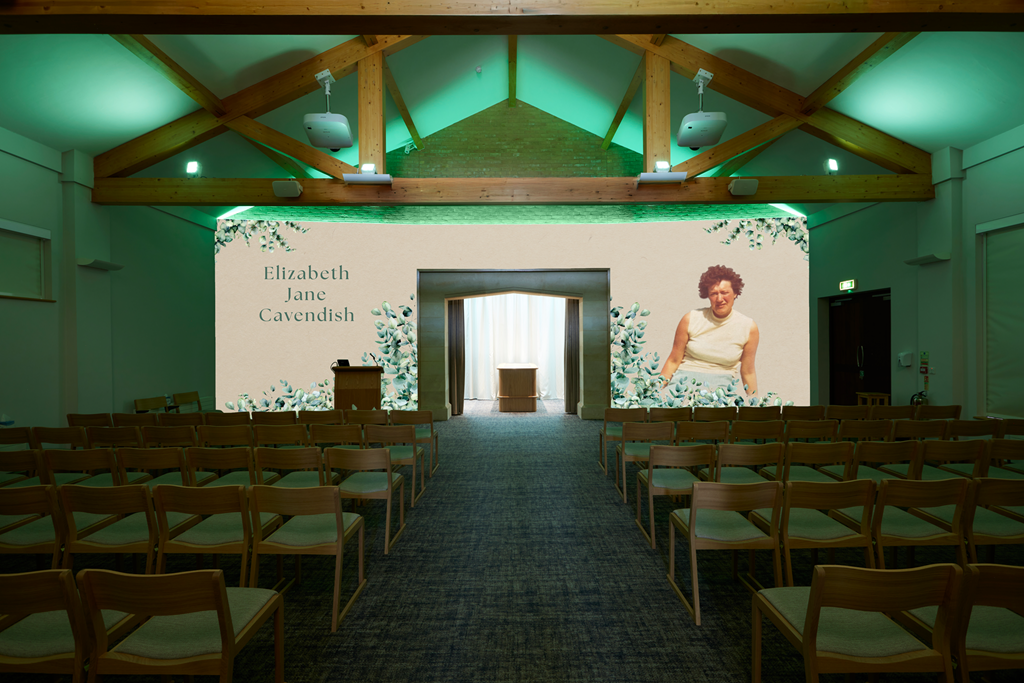 A conference or presentation room with wooden ceiling beams, rows of chairs, and a large screen displaying the name Elizabeth Jane Cavendish and a photo of a woman in a sleeveless top. There is a small podium on the left side of the screen.