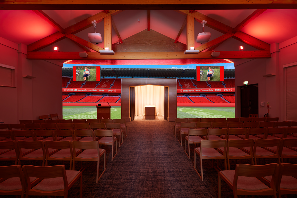 Indoor memorial or tribute space with a view of a sports stadium field outside, displaying digital screens with a photo and text honoring Elizabeth Jane Cavendish, 1946-2024, featuring a lady sitting on a bench in a park.