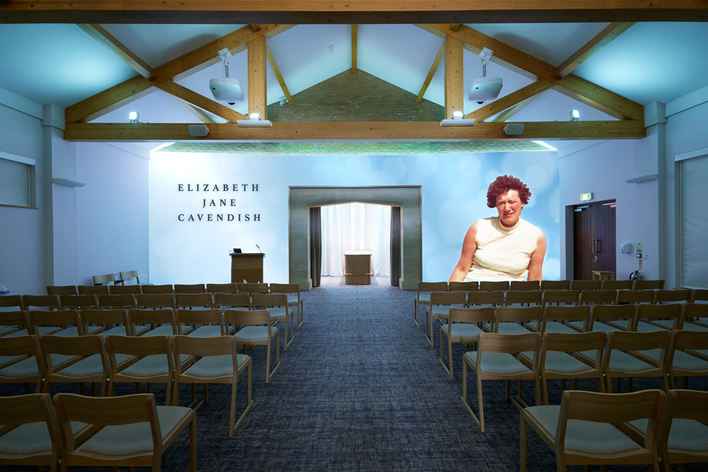 Empty auditorium with wooden chairs facing a stage, with the text 'Elizabeth Jane Cavendish' on the wall, and a large photo of a woman with curly hair on the right side.