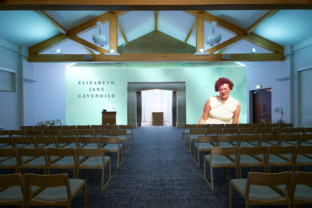 Empty auditorium with wooden chairs, a stage with a podium, and a large projected image of a woman on the right side of the stage. The background wall behind the stage displays the name Elizabeth Jane Cavendish.