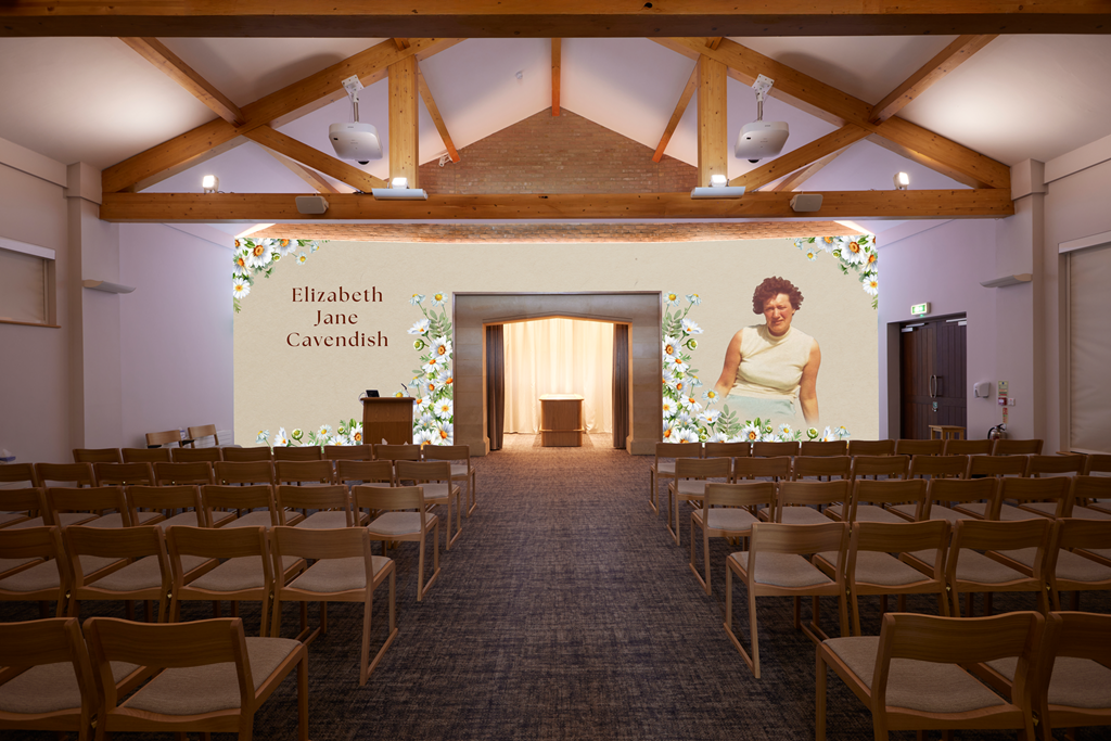 A conference or event room with rows of wooden chairs facing a large screen. The screen displays the name 'Elizabeth Jane Cavendish' surrounded by floral graphics, and a partial image of a woman in a sleeveless cream-colored top. The room has a high wooden-beamed ceiling with three mounted projectors, a podium on the stage, and a doorway at the back.
