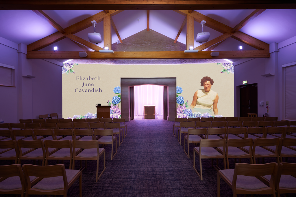 A presentation room with wooden chairs arranged in rows, a large screen displaying the names Elizabeth Jane Cavendish and a woman smiling, floral decorations around the screen, and a small stage with a lectern.