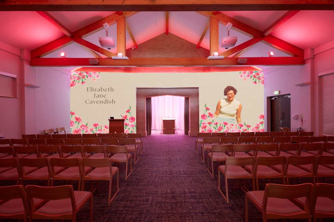 An indoor auditorium with wooden ceiling beams, pink lighting, and a large screen displaying the name Elizabeth Jane Cavendish, floral decorations, and a woman in a white dress. The room has rows of wooden chairs with cushioned seats facing the stage.